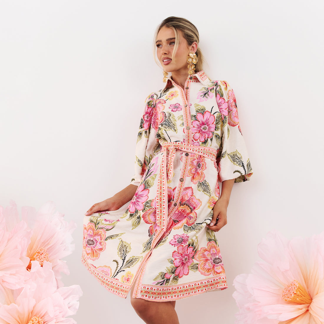 minnie + MAXXIE Lea Pink Floral Dress with gold event earrings, surrounded by pink paper flowers