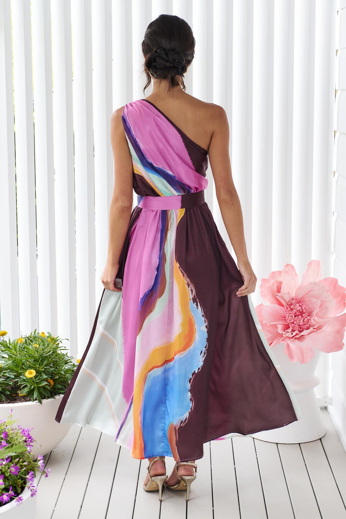 Back view of a woman wearing the minnie + MAXXIE Alessandra One Shoulder Maxi Dress with a graphic abstract print and matching waist tie.