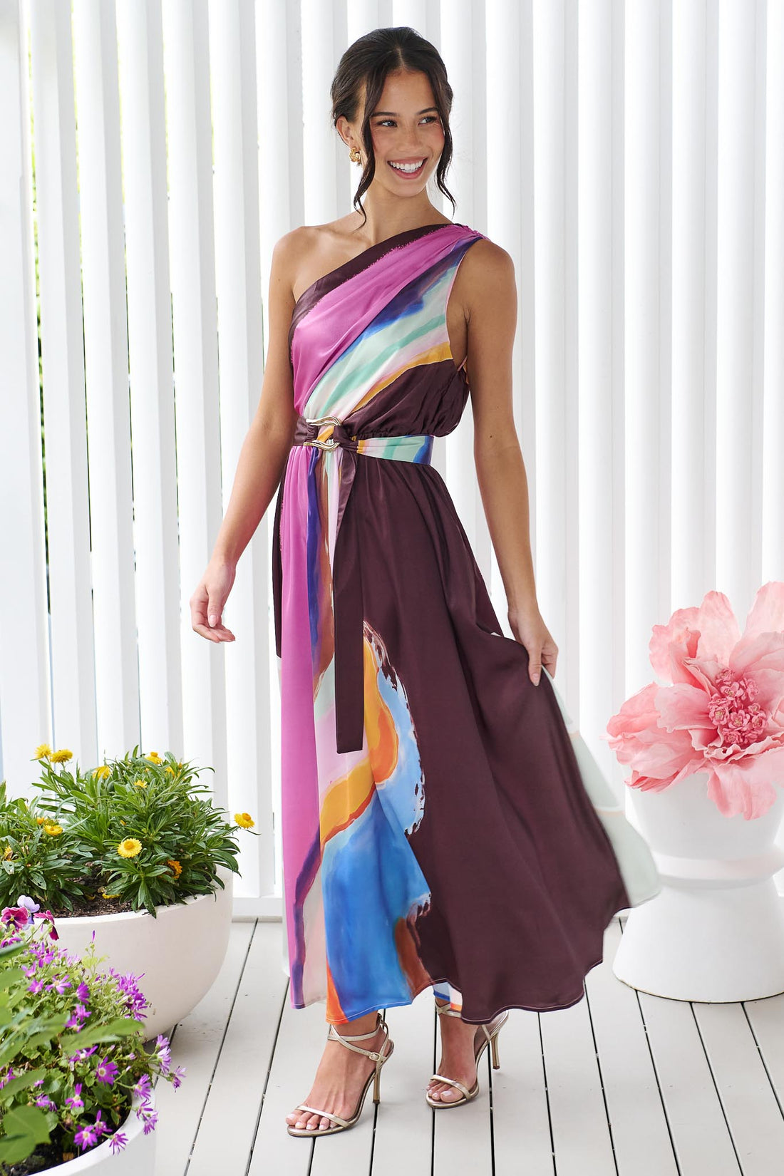 Woman wearing the minnie + MAXXIE Alessandra One Shoulder Maxi Dress with a graphic abstract print and matching waist tie.