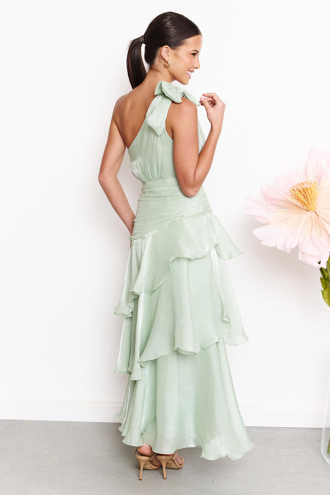 A woman wearing the minnie + MAXXIE Amara sage green one-shoulder maxi dress with a ruffled tiered skirt and shoulder bow, standing against a white wall next to a large pink flower.