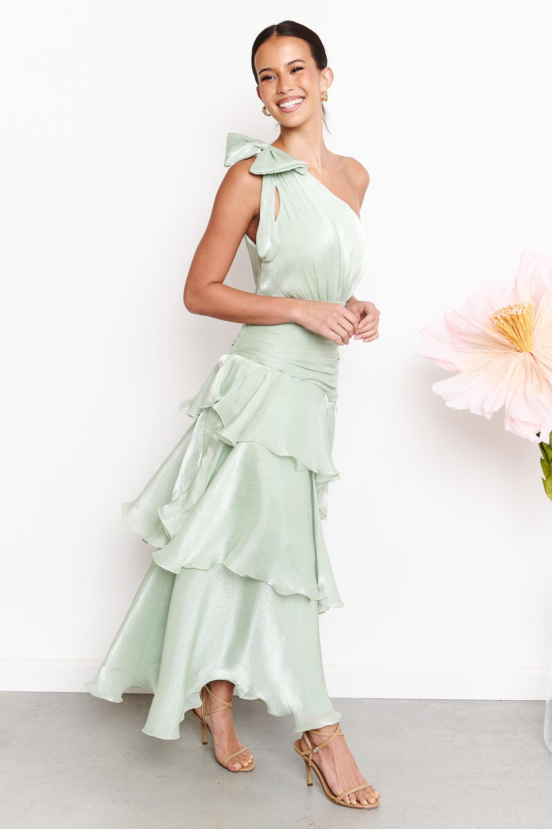 A woman wearing the minnie + MAXXIE Amara sage green one-shoulder maxi dress with a ruffled tiered skirt and shoulder bow, standing against a white wall next to a large pink flower.