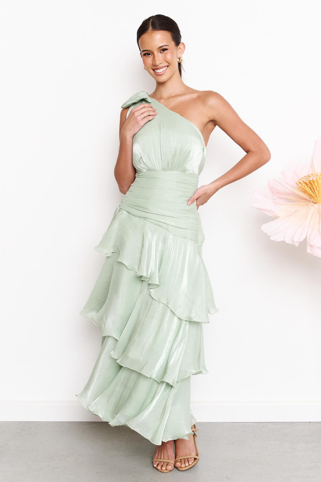 A woman wearing the minnie + MAXXIE Amara sage green one-shoulder maxi dress with a ruffled tiered skirt and shoulder bow, standing against a white wall next to a large pink flower.