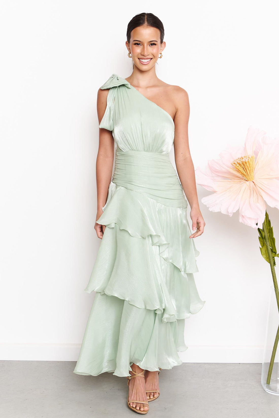A woman wearing the minnie + MAXXIE Amara sage green one-shoulder maxi dress with a ruffled tiered skirt, standing against a white wall next to a large pink flower.