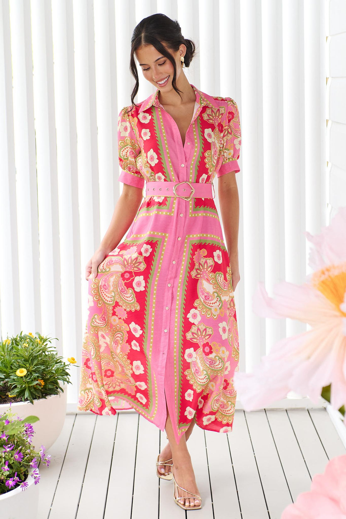 A woman wearing the minnie + MAXXIE Amaya midi shirt dress with a pink floral and paisley print and gold buckle belt, standing in front of a white panelled wall.