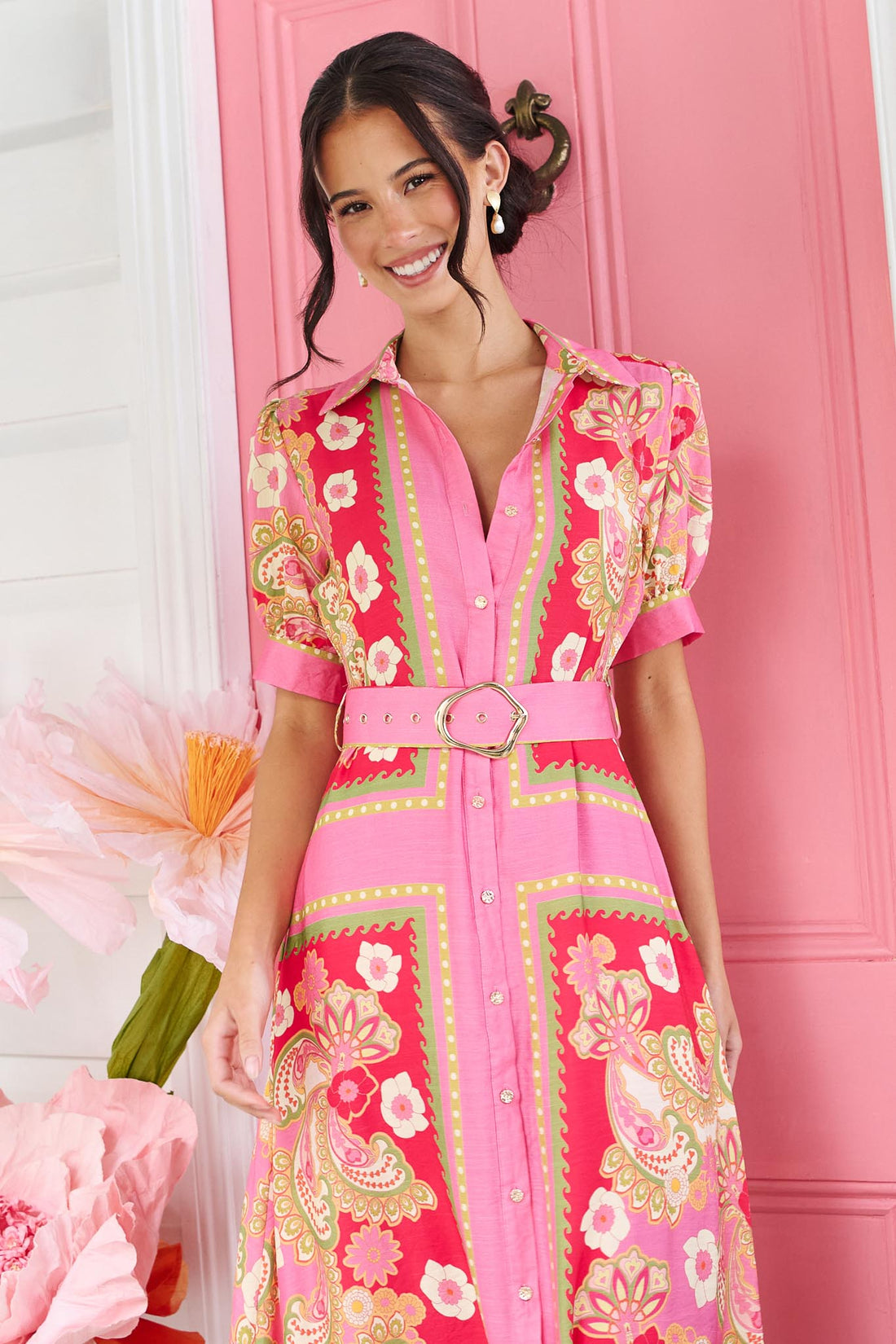 A woman wearing the minnie + MAXXIE Amaya midi shirt dress with a pink floral and paisley print and gold buckle belt, standing in front of a pink door.