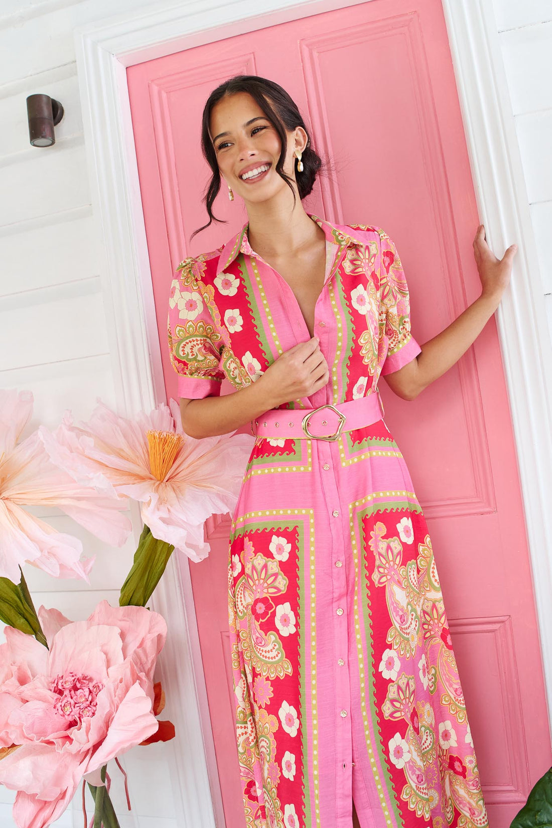 A woman wearing the minnie + MAXXIE Amaya midi shirt dress with a pink floral and paisley print and gold buckle belt, standing in front of a pink door.
