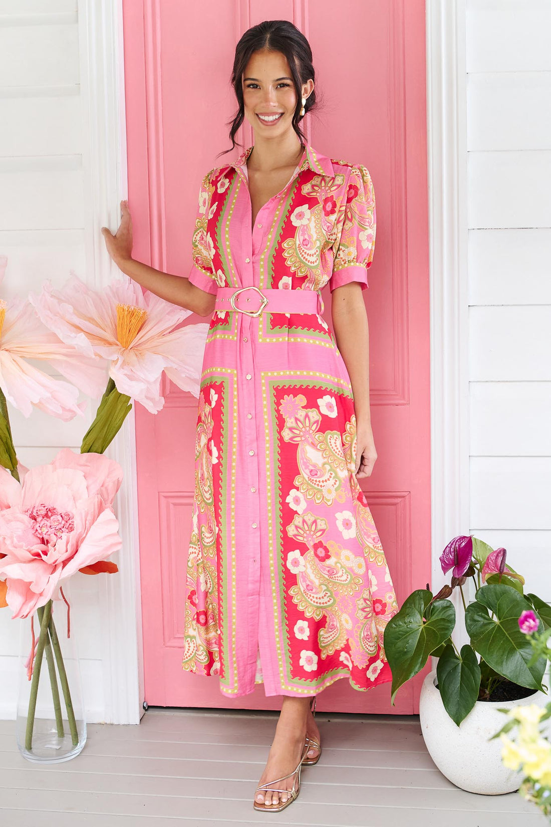 A woman wearing the minnie + MAXXIE Amaya midi shirt dress with a pink floral and paisley print and gold buckle belt, standing in front of a pink door.