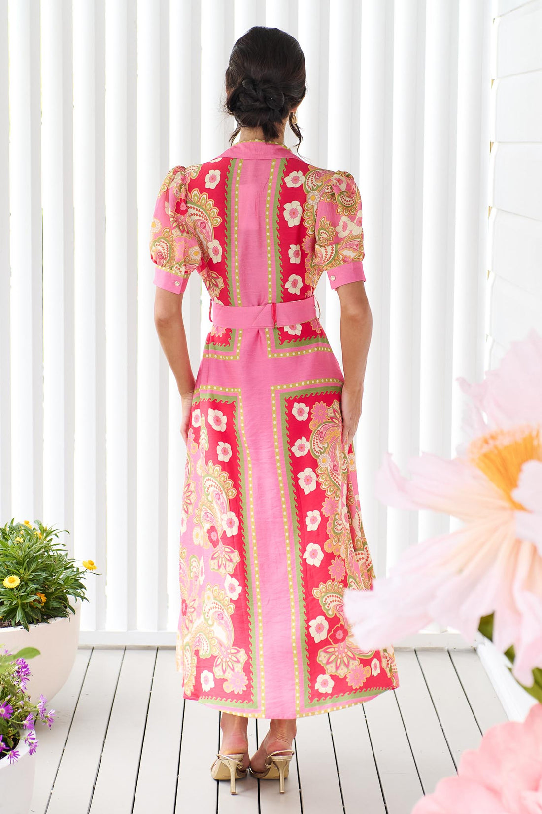 Back view of a woman wearing the minnie + MAXXIE Amaya midi shirt dress with a pink floral and paisley print and gold buckle belt, standing in front of a white panelled wall.