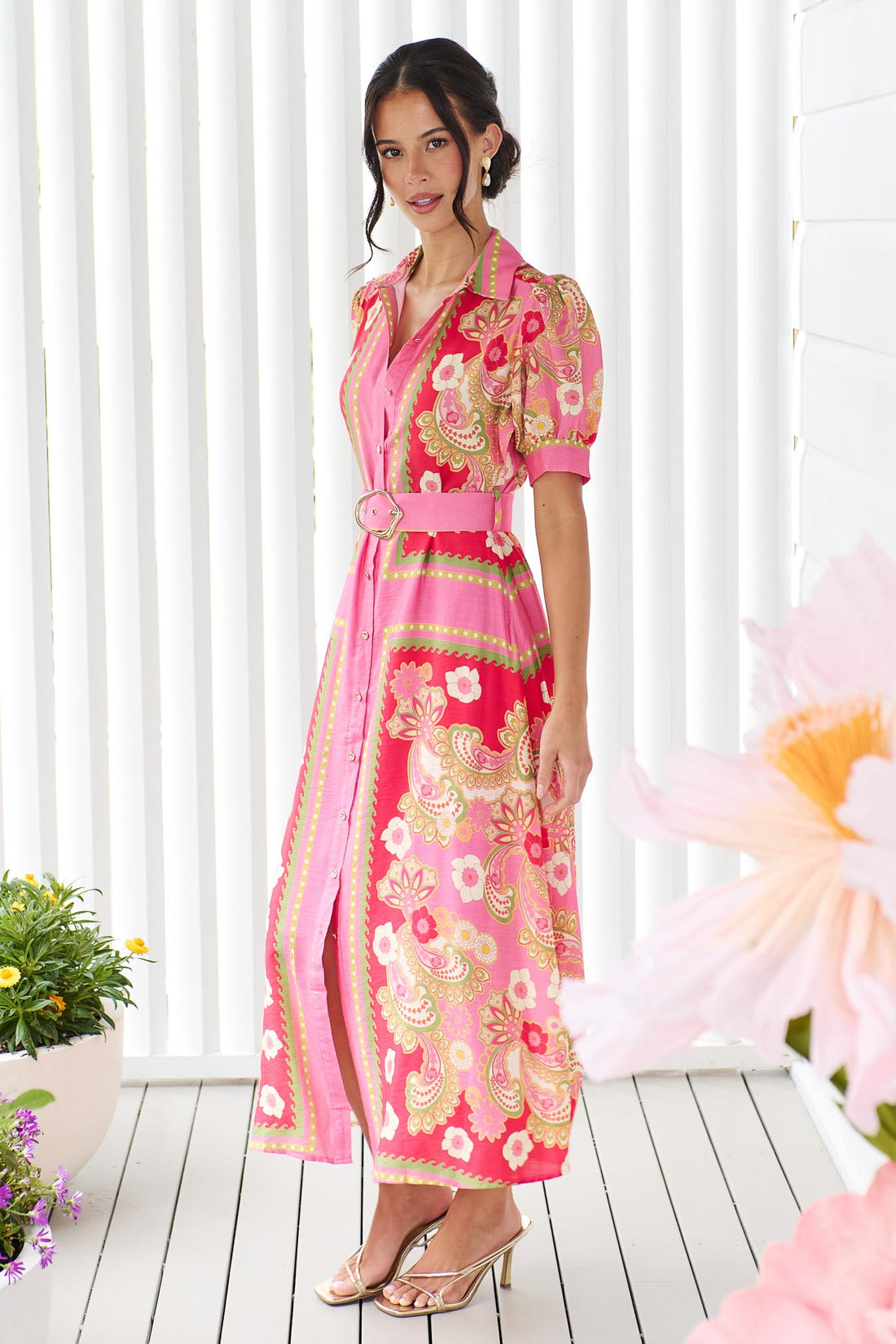 A woman wearing the minnie + MAXXIE Amaya midi shirt dress with a pink floral and paisley print and gold buckle belt, standing in front of a white panelled wall.