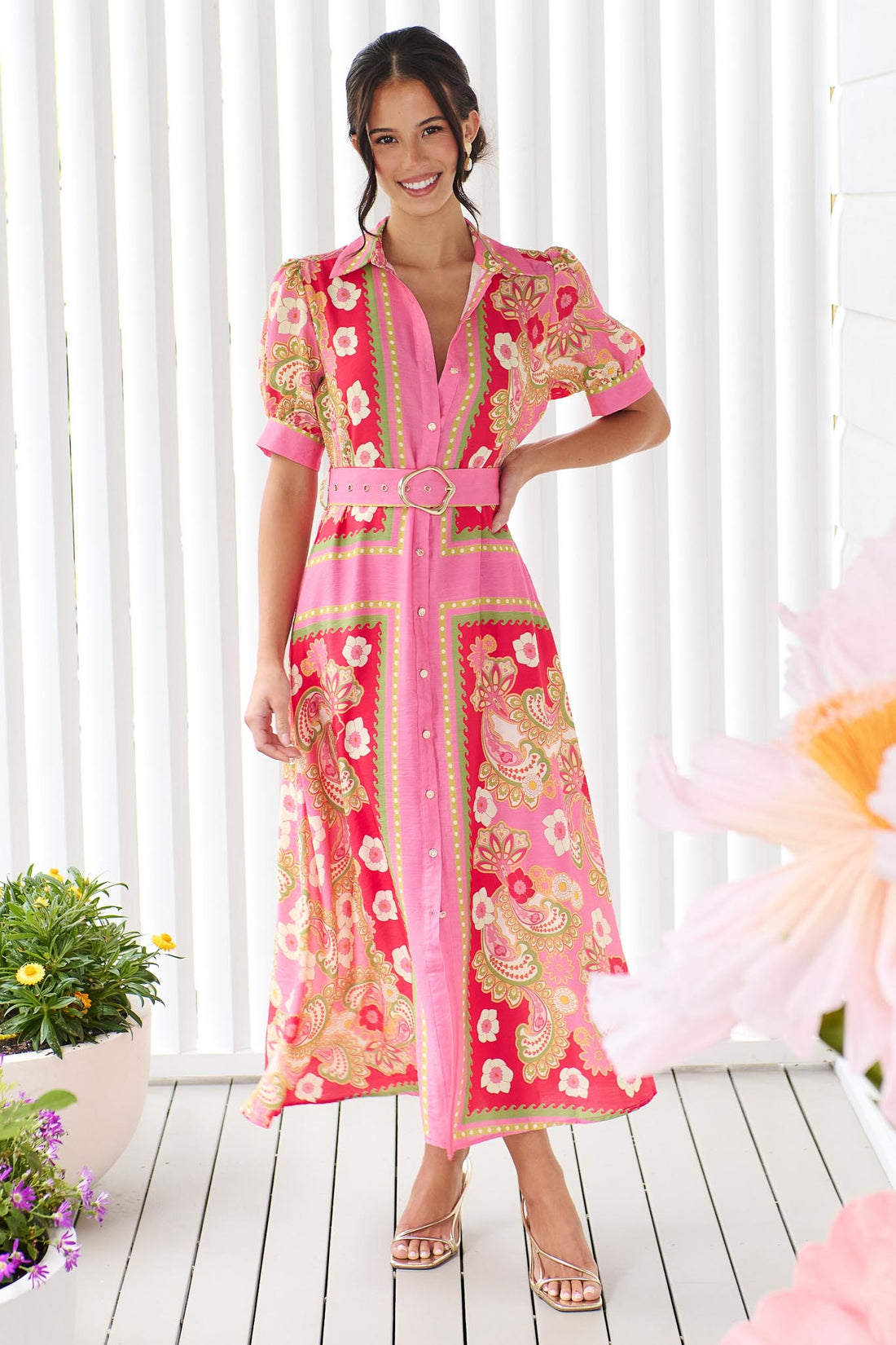 A woman wearing the minnie + MAXXIE Amaya midi shirt dress with a pink floral and paisley print and gold buckle belt, standing in front of a white panelled wall.