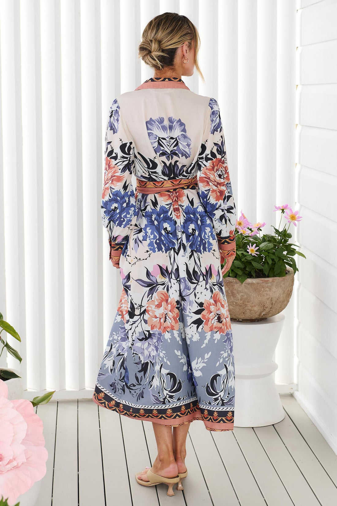 Back view of a woman wearing the minnie + MAXXIE long-sleeve floral print Arabella Midi Dress with waist tie, standing in front of a white panelled wall.