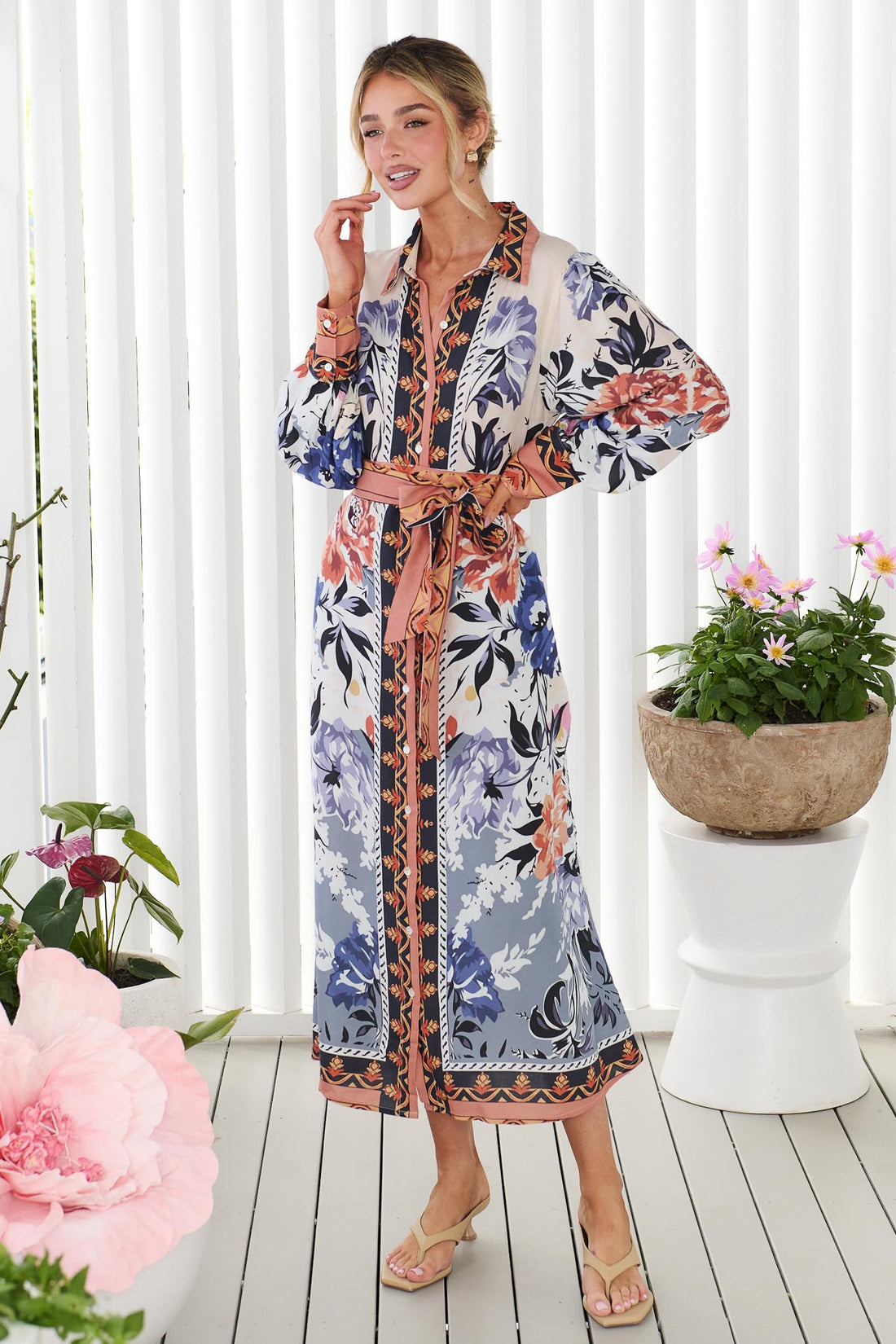 Side view of a woman wearing the minnie + MAXXIE long-sleeve floral print Arabella Midi Dress with waist tie, standing in front of a white panelled wall with potted flowers.