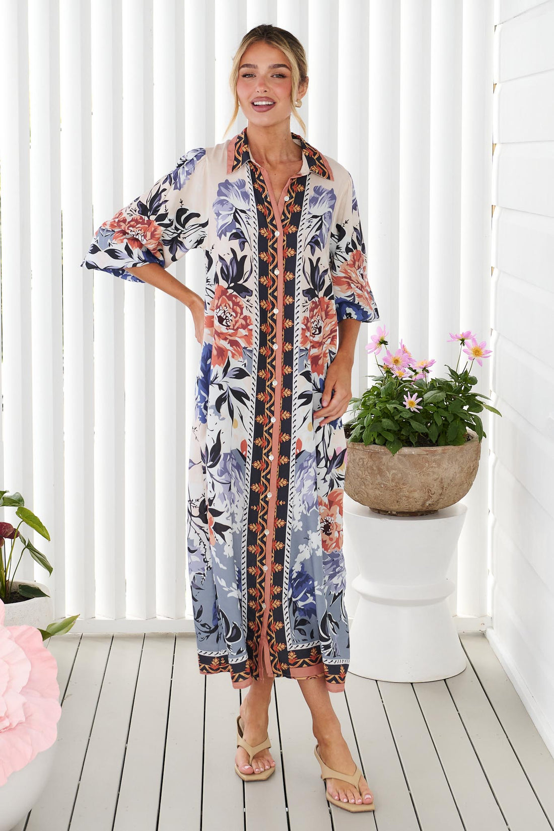 A woman wearing the minnie + MAXXIE long-sleeve floral print Arabella Midi Dress, standing in front of a white panelled wall with potted flowers.