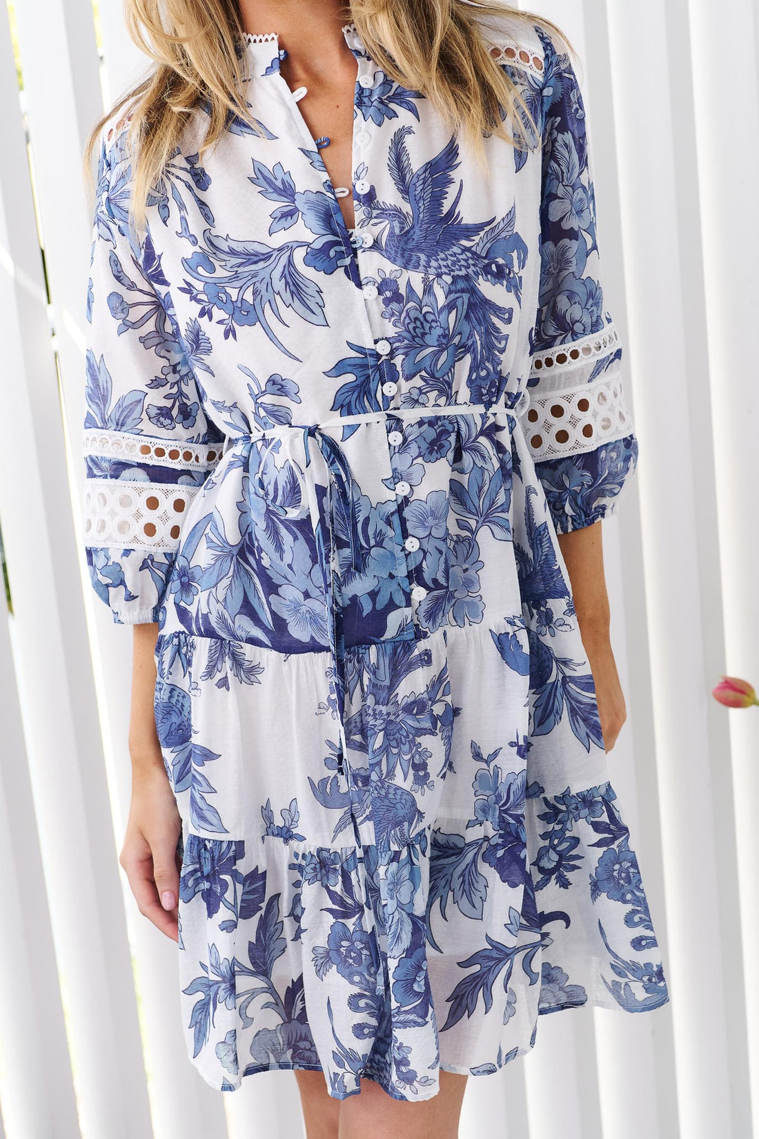 Detail of a woman wearing the minnie + MAXXIE Arde blue and white floral print dress with a tie waist and circle lace sleeves, standing in front of a white panelled wall.