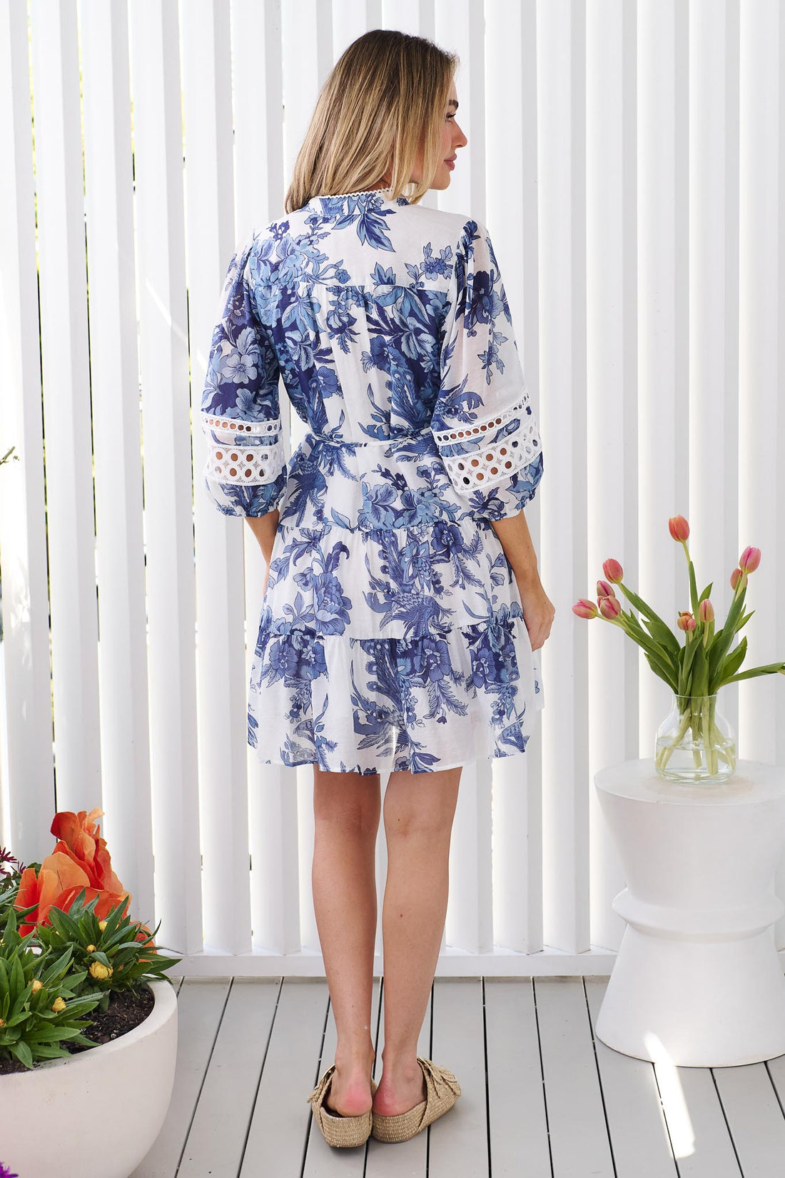 Back view of a woman wearing the minnie + MAXXIE Arde blue and white floral print dress with a tie waist and circle lace sleeves, standing in front of a white panelled wall.
