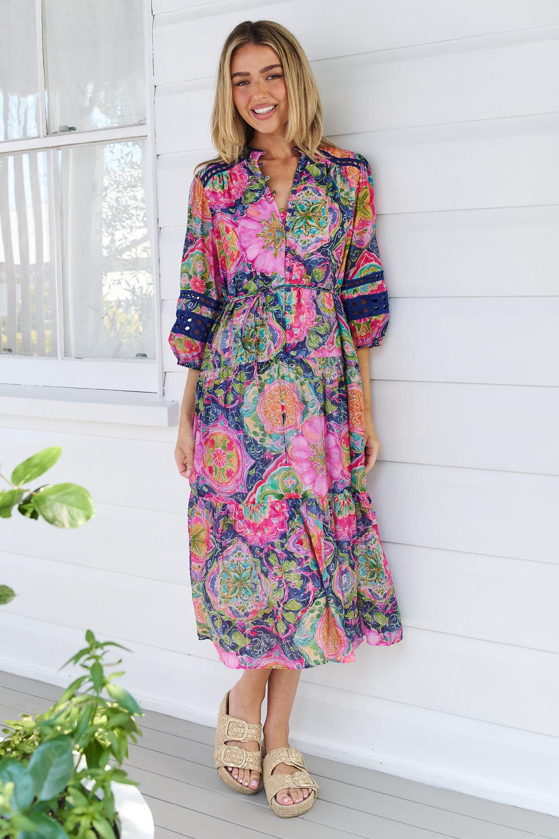 A woman wearing the minnie + MAXXIE Arden Midi Dress in watercolour floral with blue lace inserts, standing in front of a white wall.