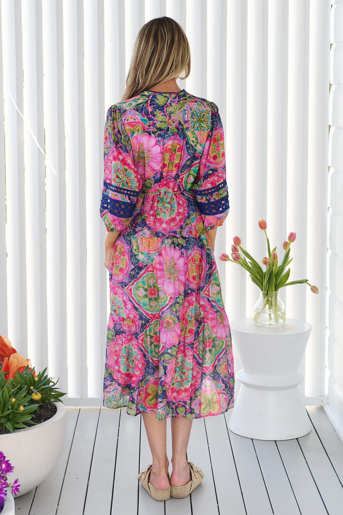 Back view of a woman wearing the minnie + MAXXIE Arden Midi Dress in watercolour floral with blue lace inserts, standing in front of a white panelled wall.