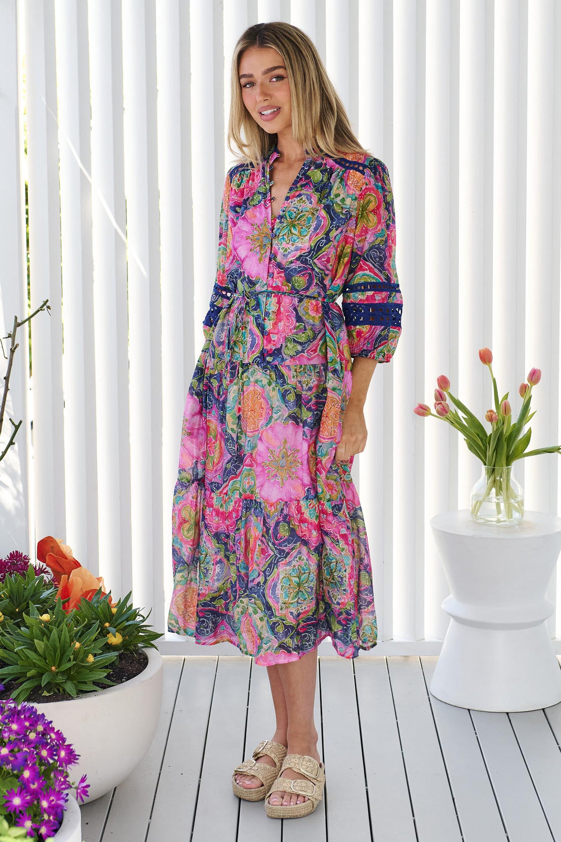 Side view of a woman wearing the minnie + MAXXIE Arden Midi Dress in watercolour floral with blue lace inserts, standing in front of a white panelled wall.