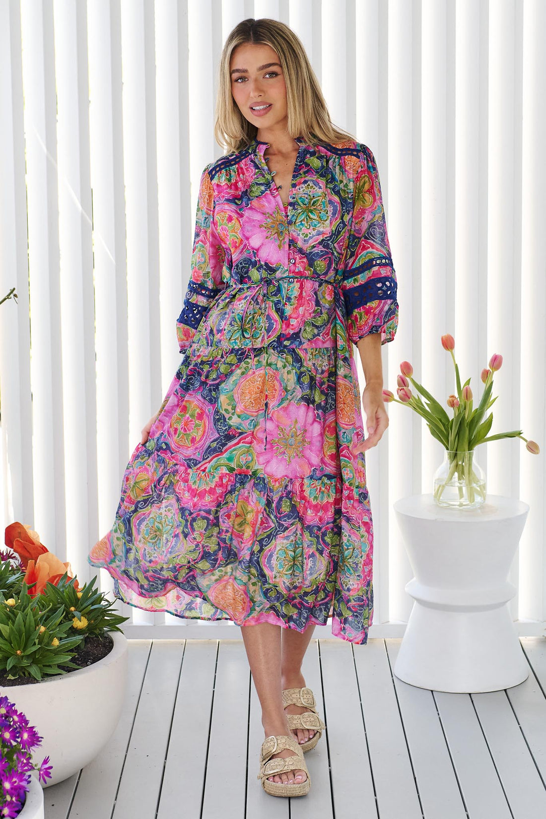 A woman wearing the minnie + MAXXIE Arden Midi Dress in watercolour floral with blue lace inserts, standing in front of a white panelled wall.