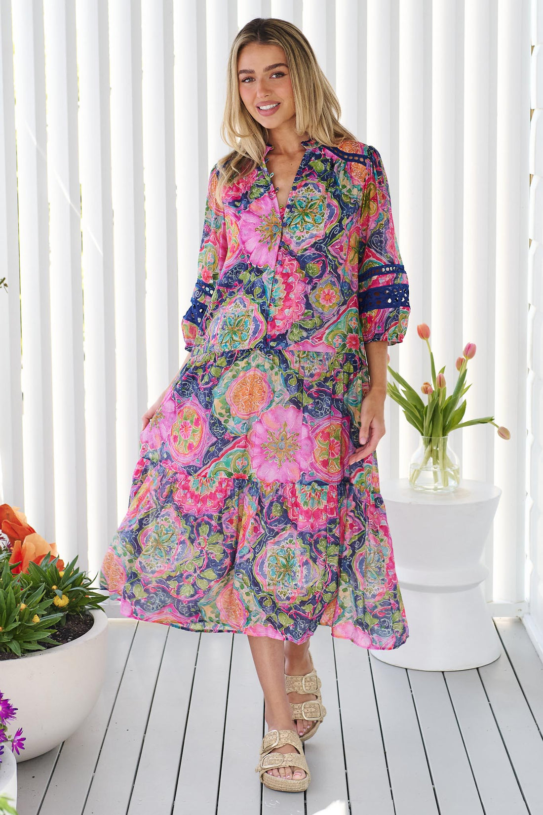 A woman wearing the minnie + MAXXIE Arden Midi Dress in watercolour floral with blue lace inserts, standing in front of a white panelled wall.