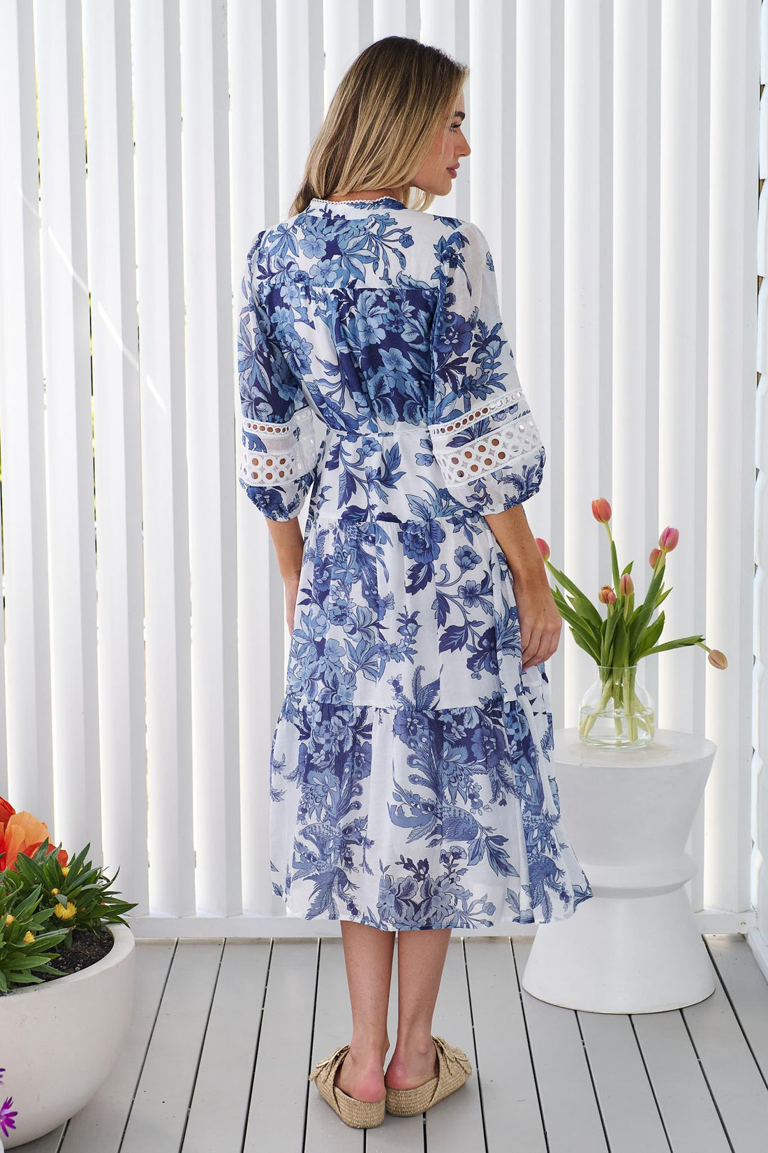 Back view of a woman wearing the minnie + MAXXIE Arden blue and white floral print midi dress with a tie waist and circle lace details, standing in front of a white panelled wall.