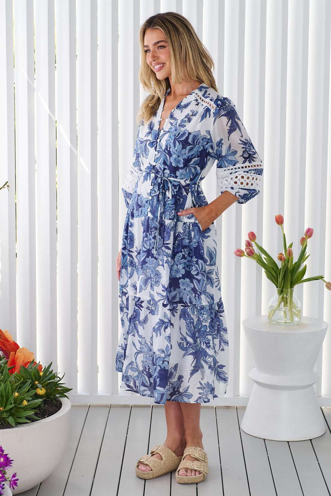 Side view of a woman wearing the minnie + MAXXIE Arden blue and white floral print midi dress with a tie waist and circle lace details, standing in front of a white panelled wall.