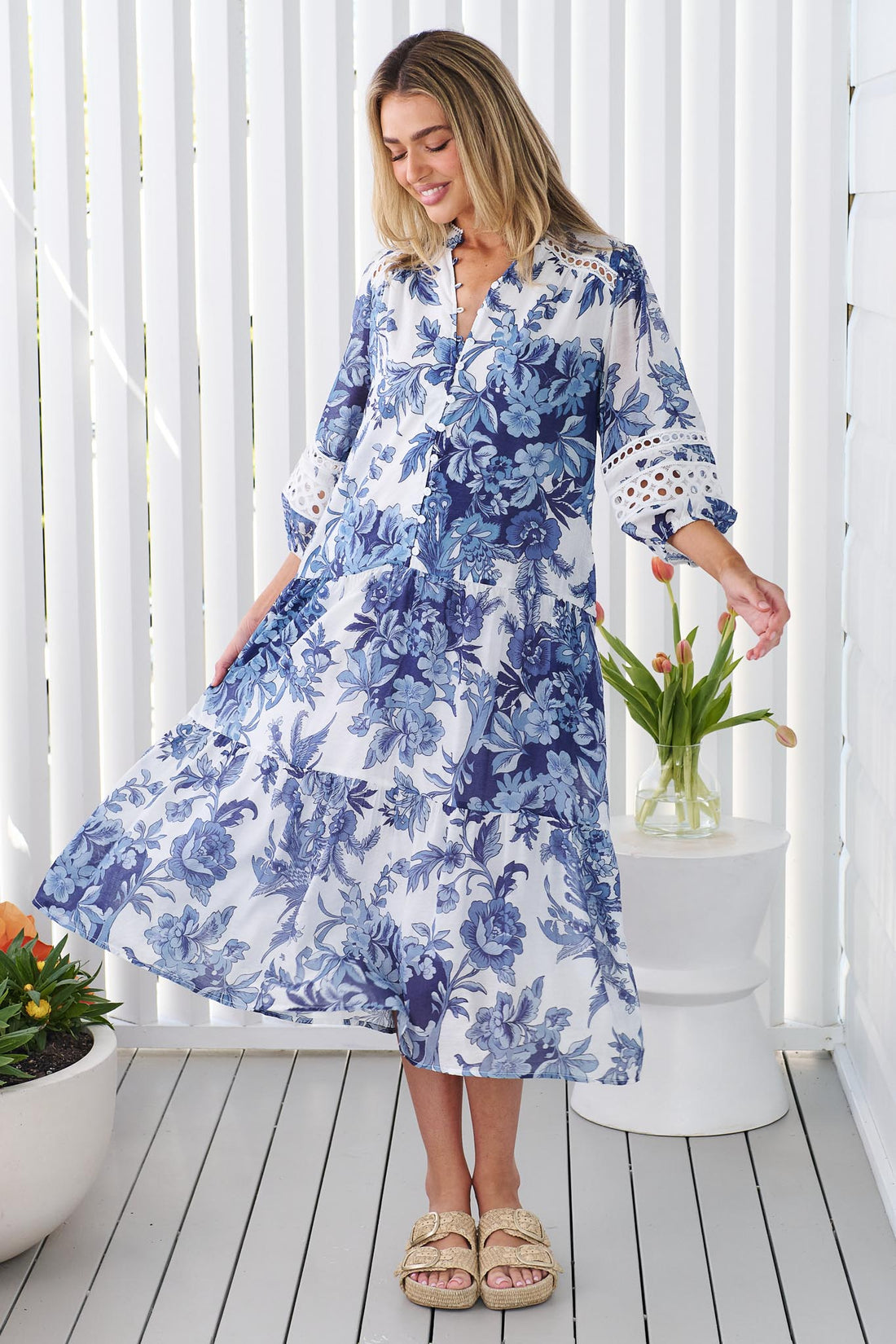 A woman wearing the minnie + MAXXIE Arden blue and white floral print midi dress with circle lace details, standing in front of a white panelled wall.