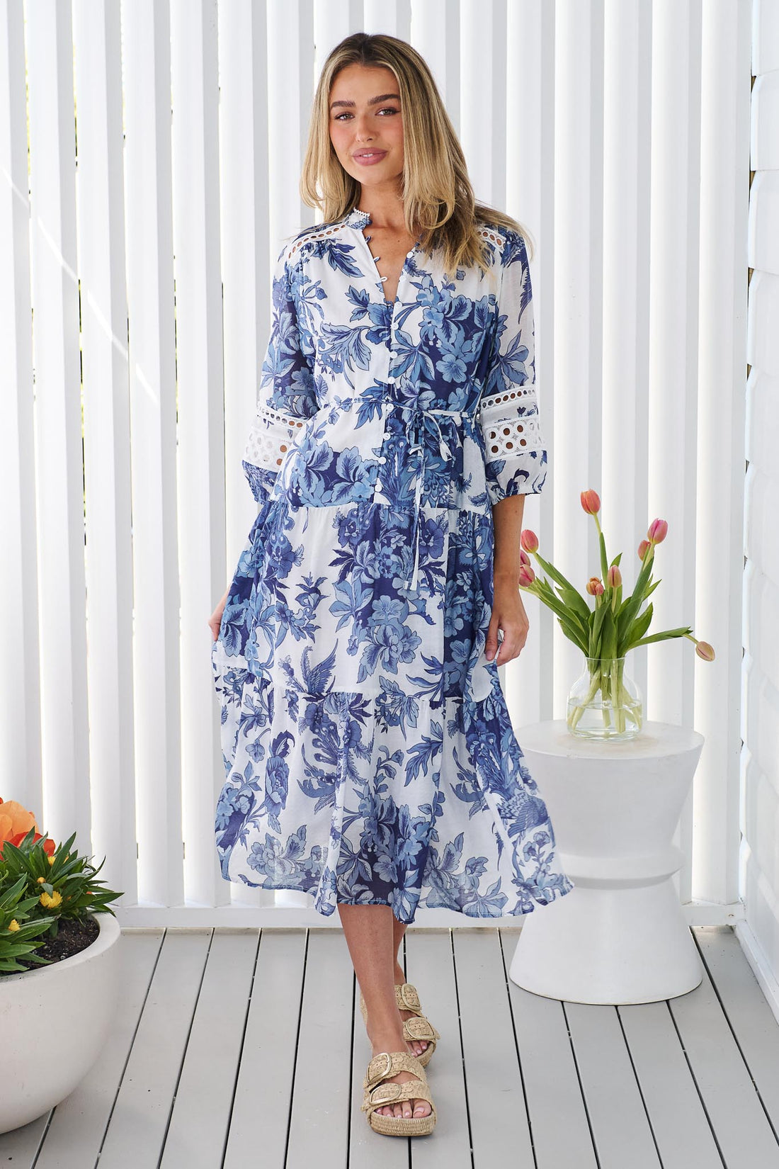 A woman wearing the minnie + MAXXIE Arden blue and white floral print midi dress with a tie waist and circle lace details, standing in front of a white panelled wall.