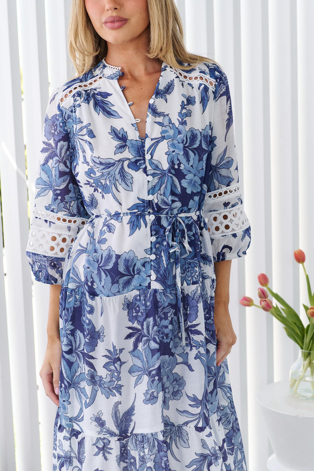 Detail of a woman wearing the minnie + MAXXIE Arden blue and white floral print midi dress with a tie waist and circle lace details, standing in front of a white panelled wall.