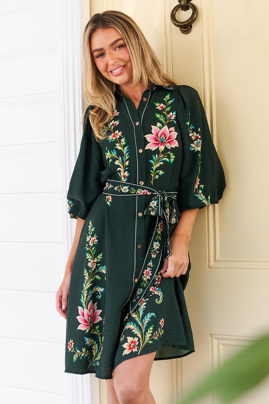 A woman wearing the minnie + MAXXIE dark green Arielle shirt dress with pink and green floral print, standing in front of a yellow door.