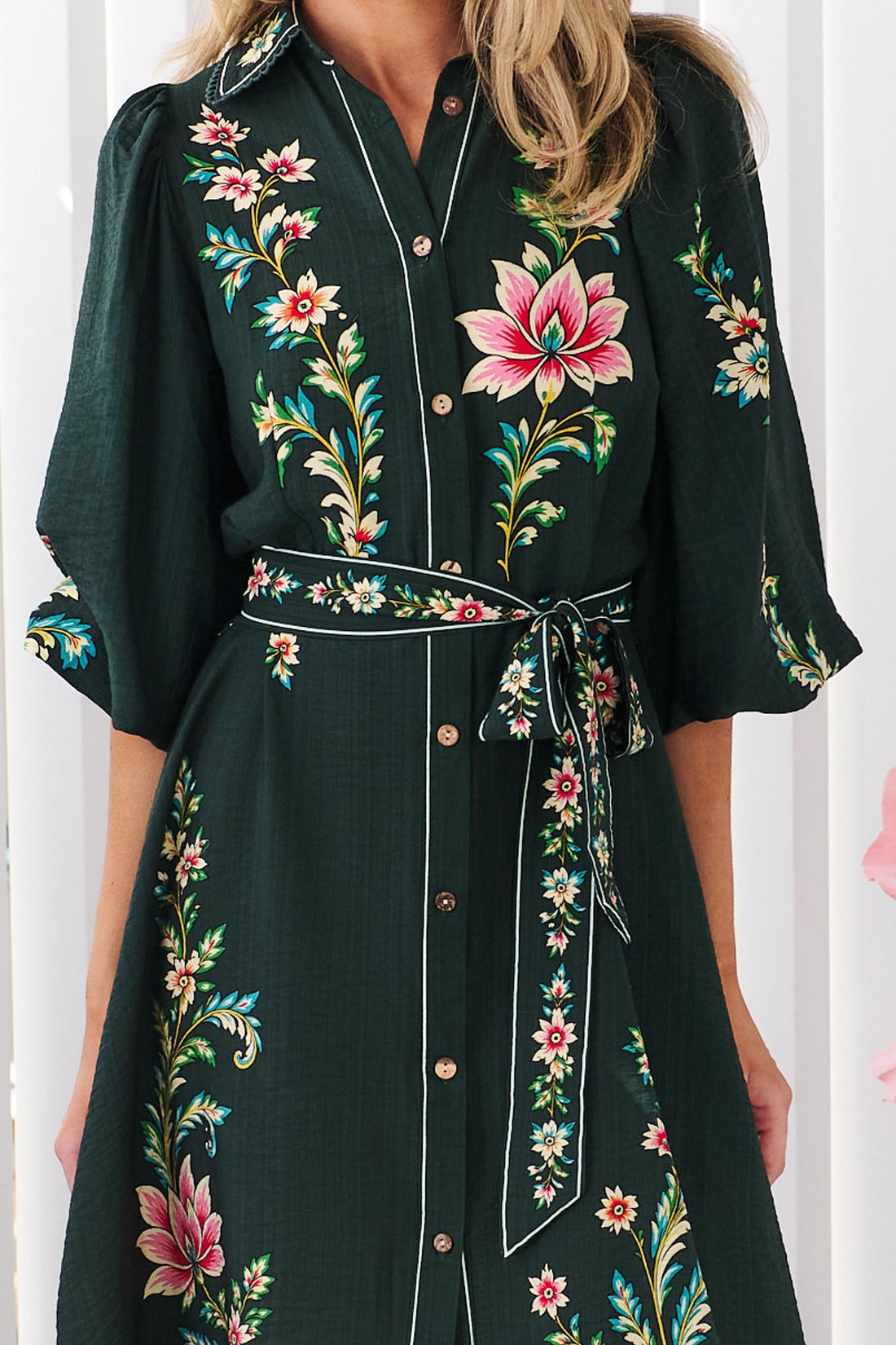 Close up of a woman wearing the minnie + MAXXIE dark green Arielle shirt dress with pink and green floral print, standing in front of a white panelled wall.