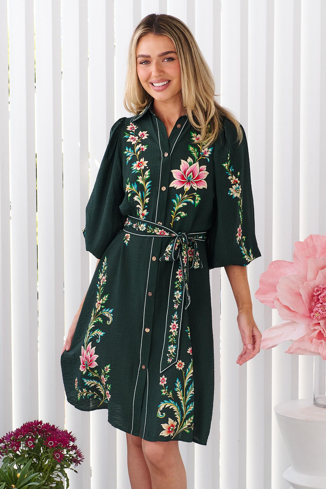 A woman wearing the minnie + MAXXIE dark green Arielle shirt dress with pink and green floral print, standing in front of a white panelled wall.