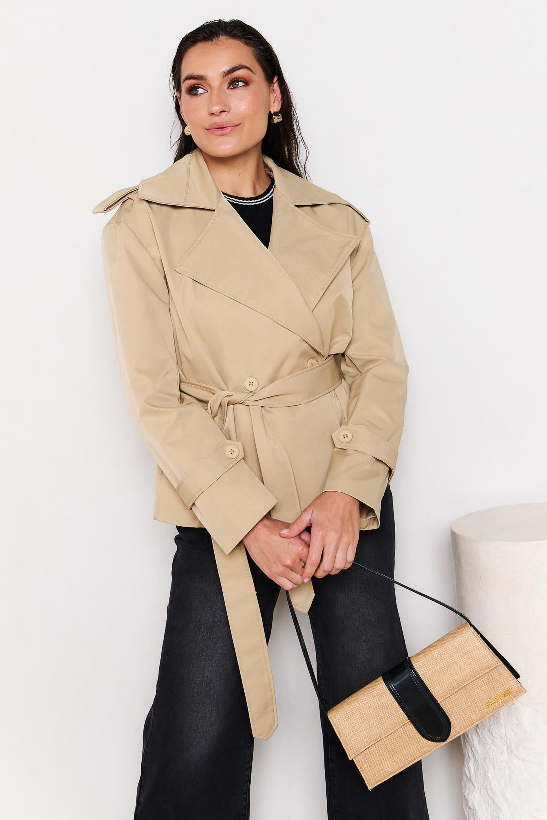 Woman wearing a minnie + MAXXIE beige cropped trench coat holding a matching jacquemus handbag against a white background