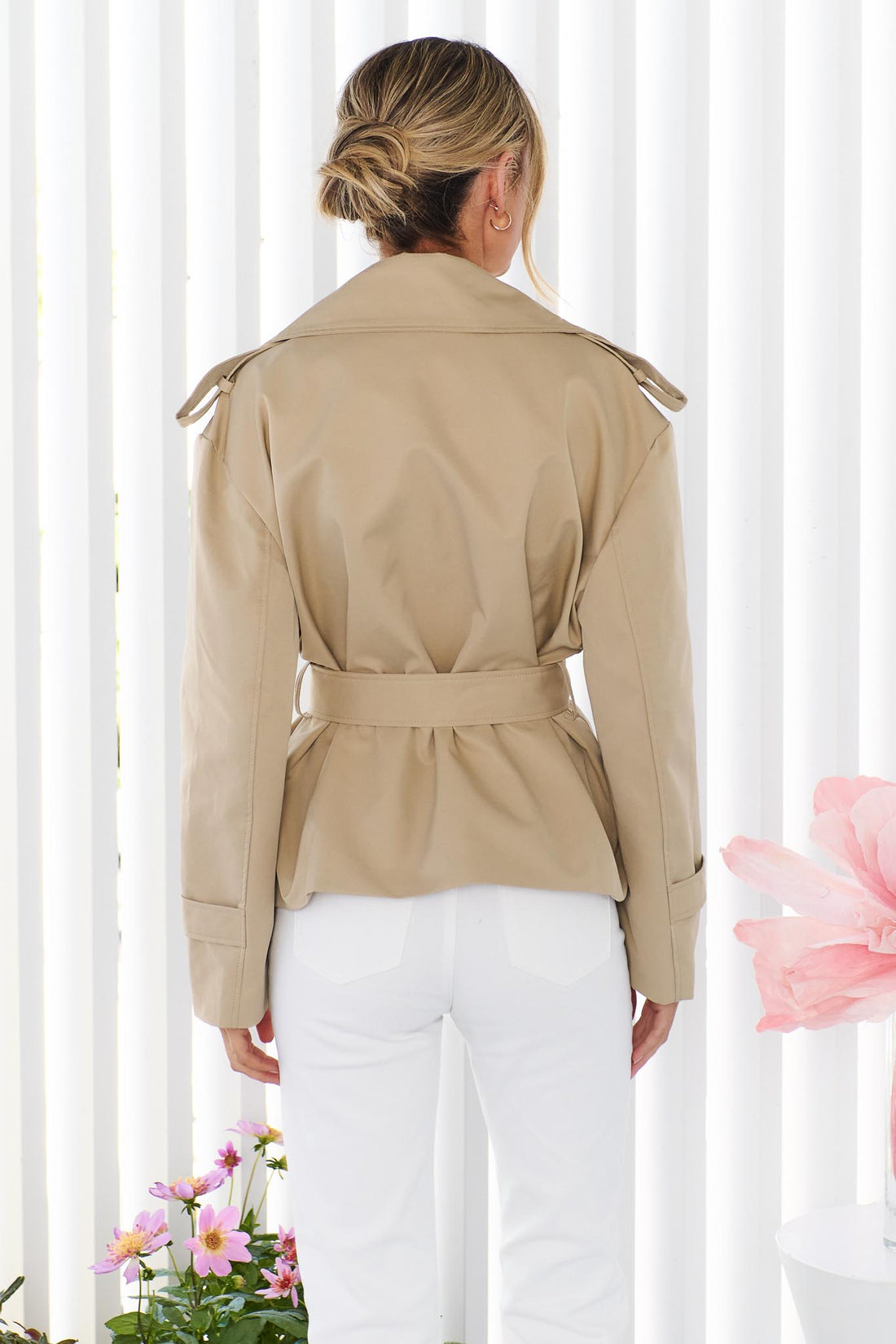 A woman viewed from the back wearing a beige belted trench coat and white pants, standing in front of white vertical wall panels with pink flowers at the bottom corners.