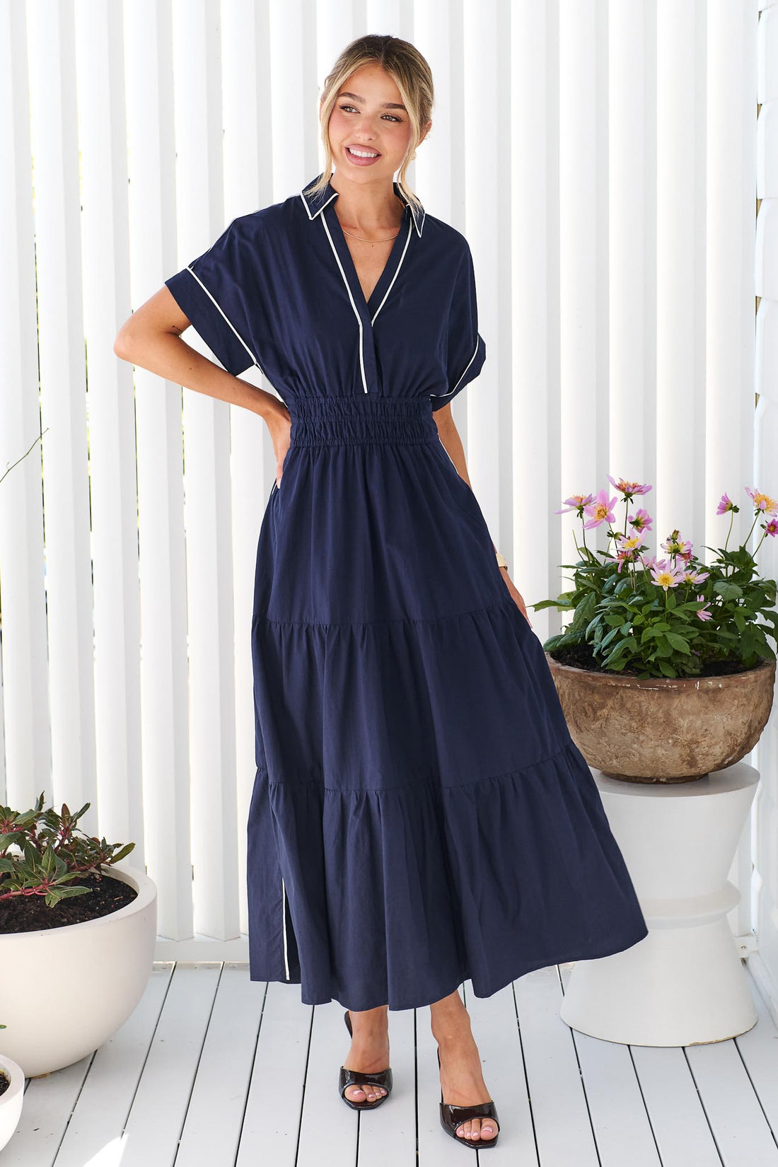 A woman wearing the minnie + MAXXIE Audrey navy tiered midi dress with white piping details, standing on a white deck in front of a white slatted wall with potted plants.