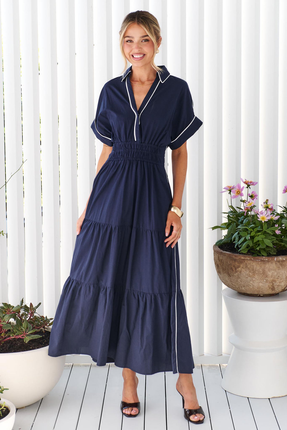A woman wearing the minnie + MAXXIE Audrey navy tiered maxi dress with white piping details, standing on a white deck in front of a white slatted wall with potted plants.
