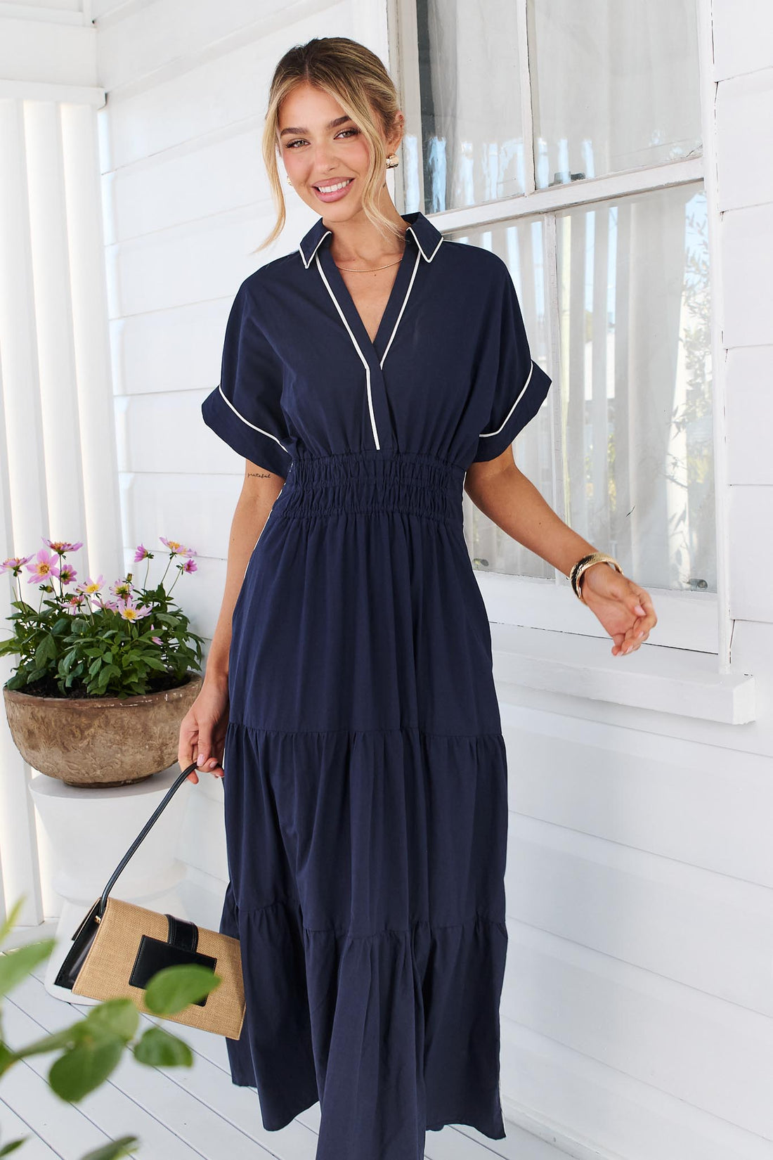 A woman wearing the minnie + MAXXIE Audrey navy tiered midi dress with white piping details, standing on a white deck in front of a white slatted wall with potted plants.