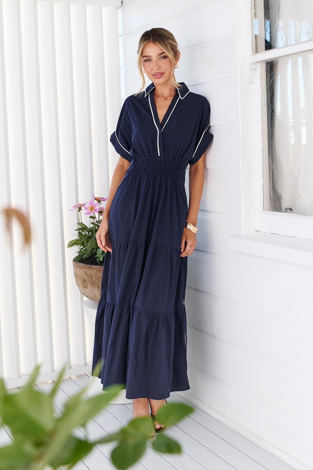 A woman wearing the minnie + MAXXIE Audrey navy tiered midi dress with white piping details, standing on a white deck in front of a white slatted wall with potted plants.