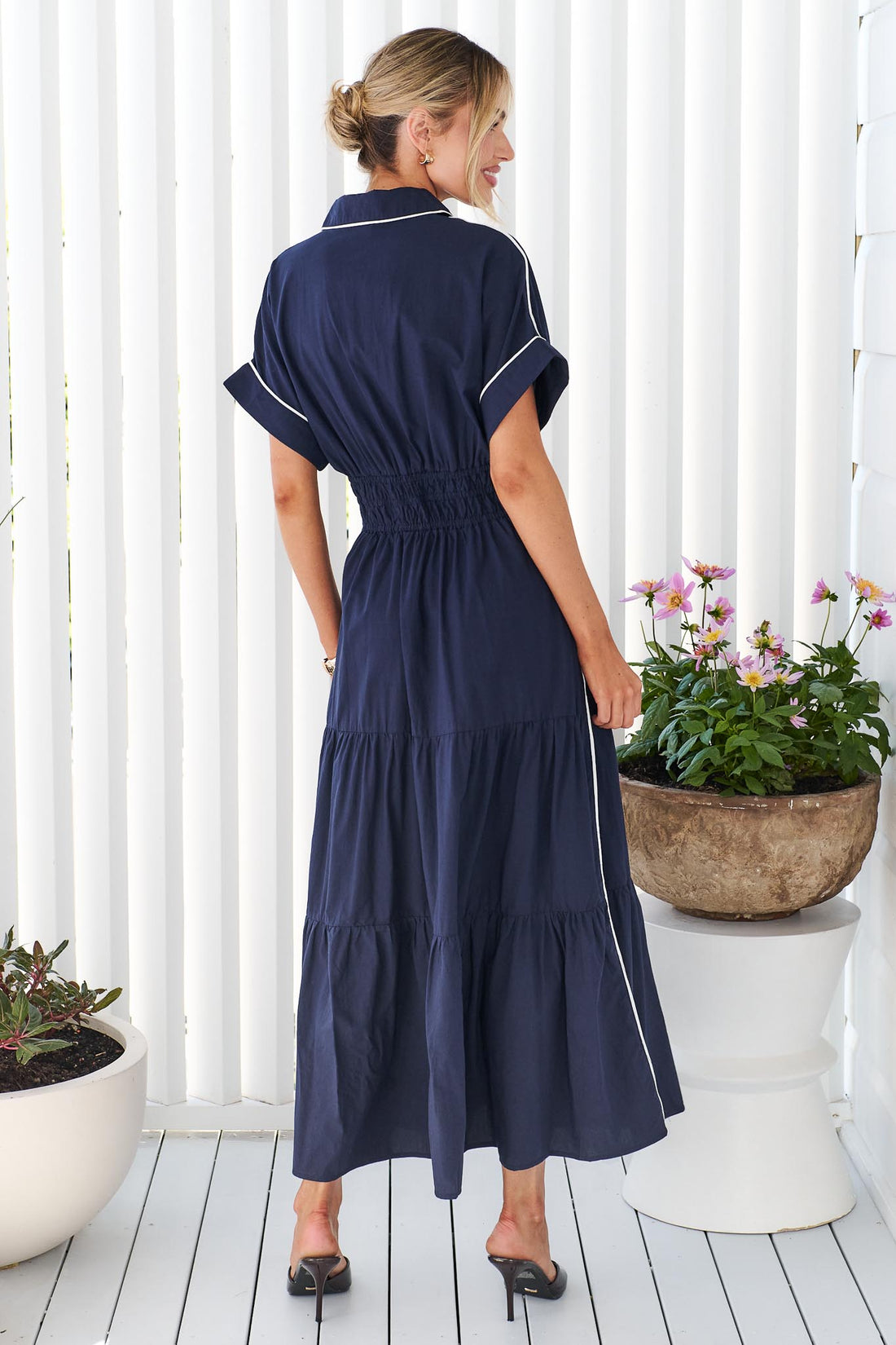 A woman wearing the minnie + MAXXIE Audrey navy tiered midi dress with white piping details, standing on a white deck in front of a white slatted wall with potted plants.