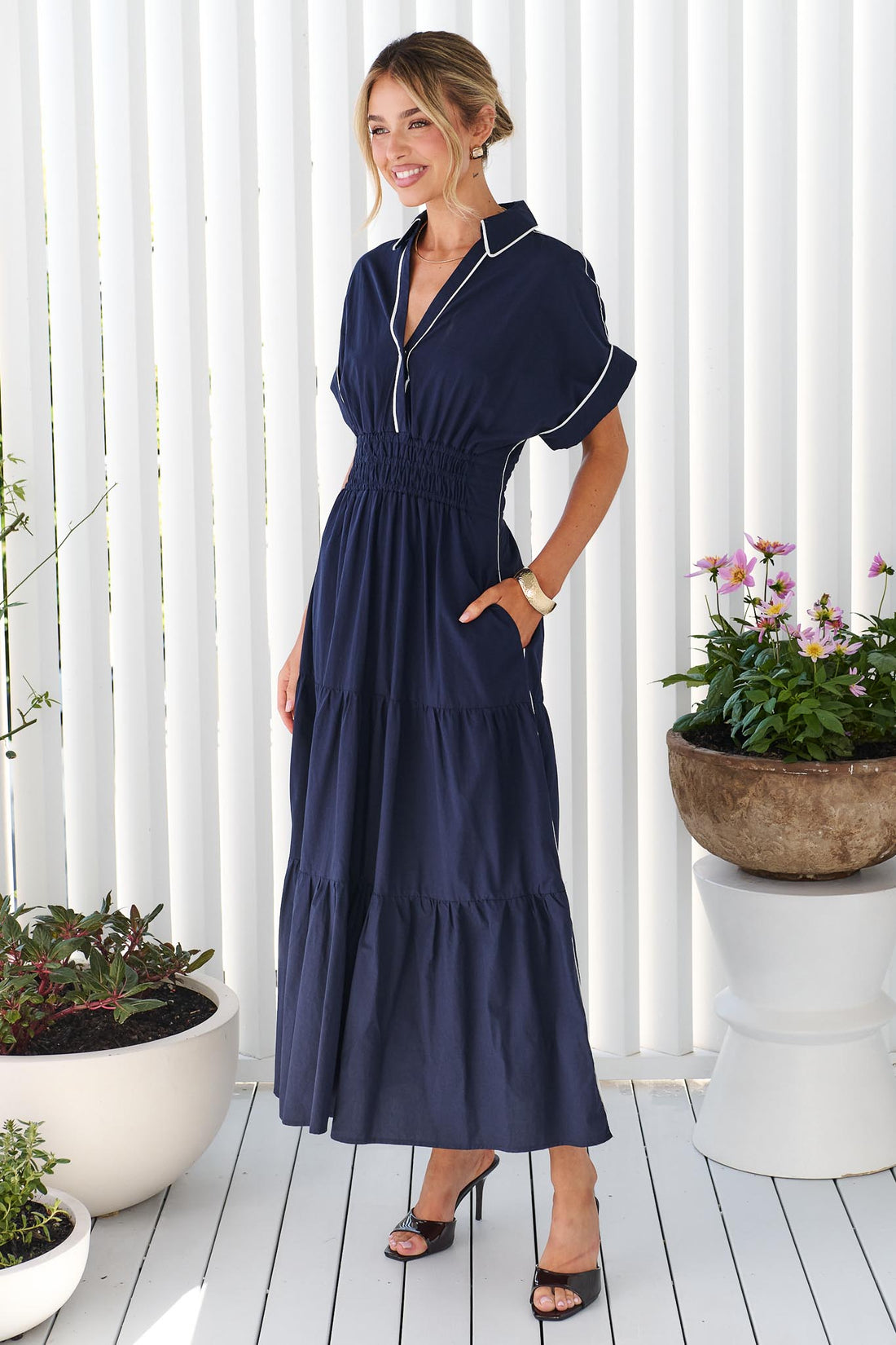 A woman wearing the minnie + MAXXIE Audrey navy tiered midi dress with white piping details, standing on a white deck in front of a white slatted wall with potted plants.