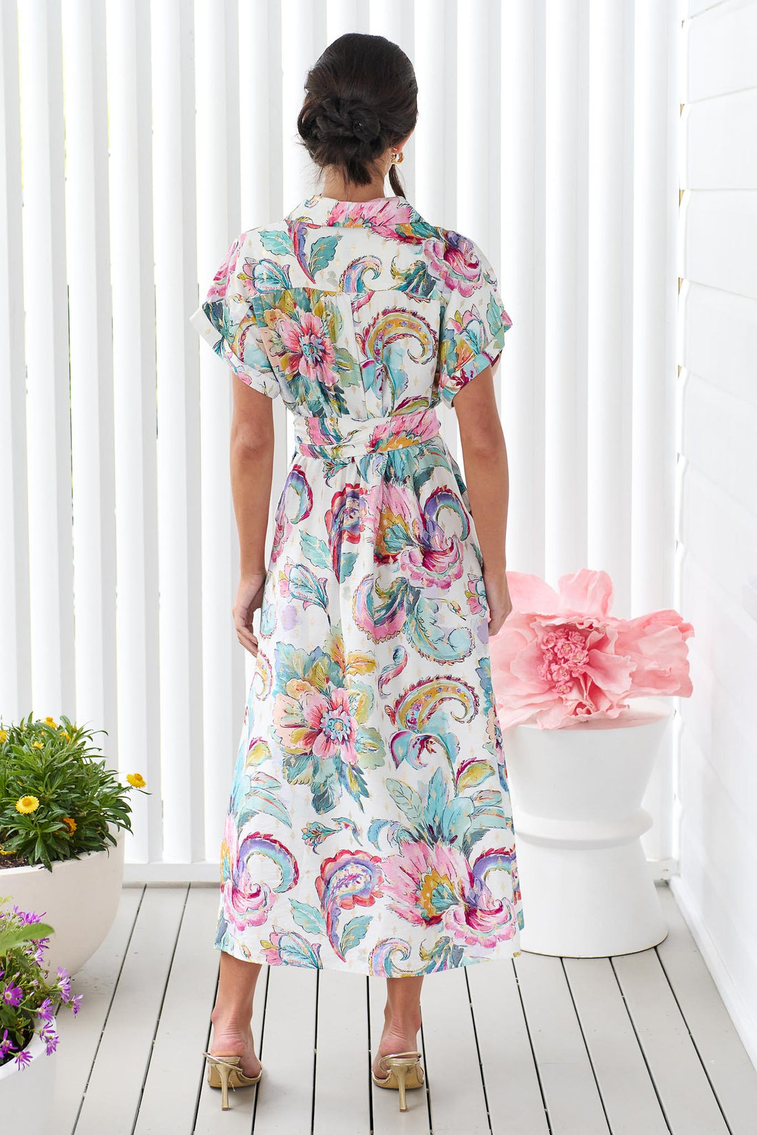 Back view of a woman wearing the minnie + MAXXIE short-sleeve Audrey midi dress with a colourful floral and paisley print and matching waist tie, standing in front of a white panelled wall.