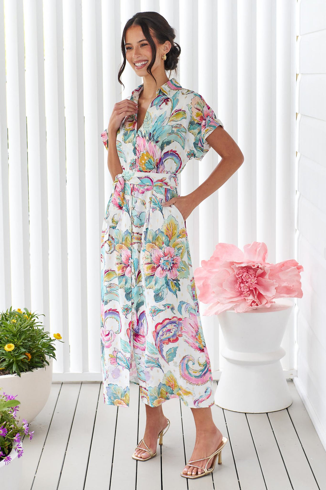  A woman wearing the minnie + MAXXIE short-sleeve Audrey midi dress with a colourful floral and paisley print and matching waist tie, standing in front of a white panelled wall.