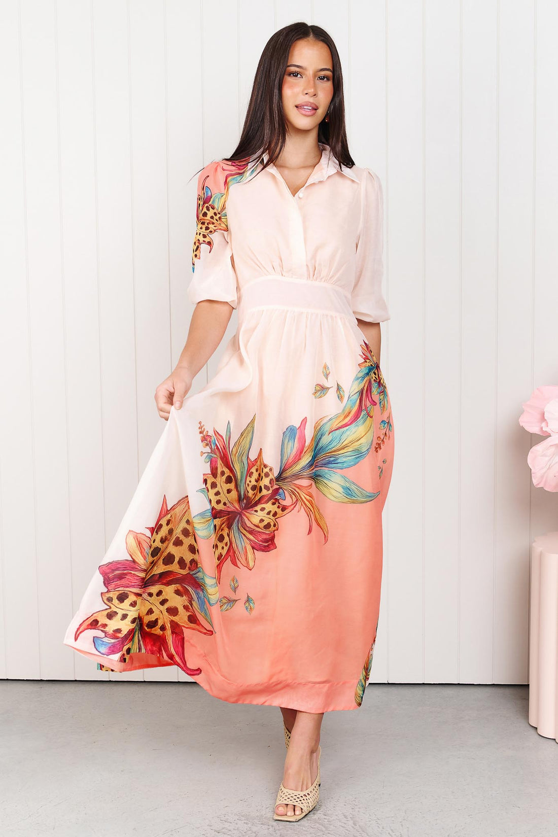 Woman wearing the minnie + MAXXIE Ayla Havana midi event dress in soft peach and pink with a leopard floral print, standing in front of a white panelled wall.