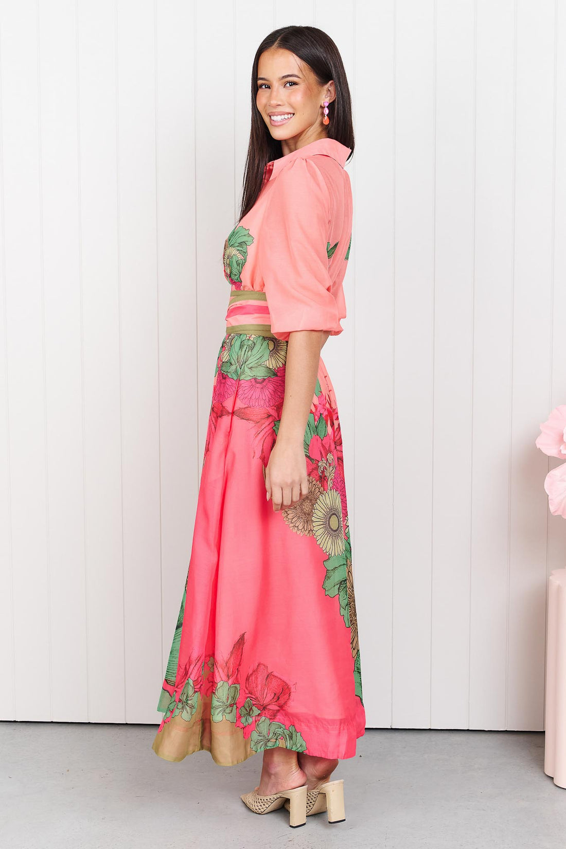 Woman wearing the minnie + MAXXIE Ayla Madison Ave midi event dress in peach and pink with a floral print and stripe waist tie, standing in front of a white panelled wall.