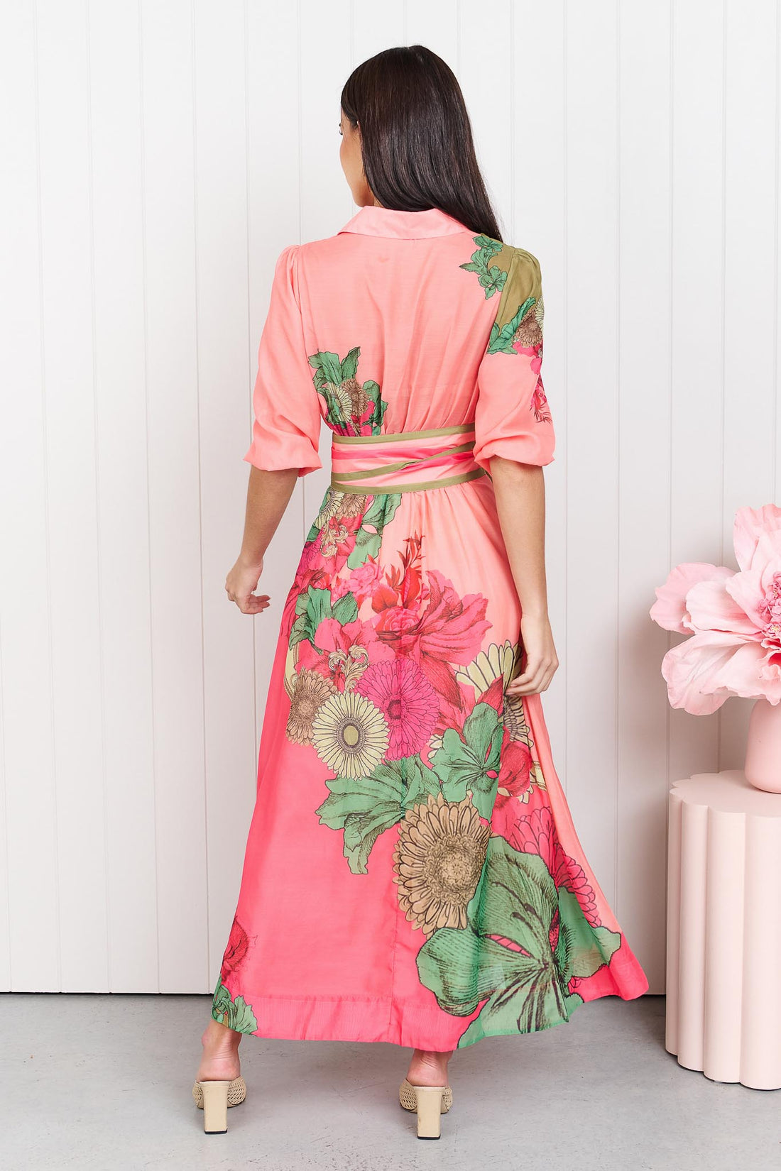 Back view of a woman wearing the minnie + MAXXIE Ayla Madison Ave midi event dress in peach and pink with a floral print and stripe waist tie, standing in front of a white panelled wall.