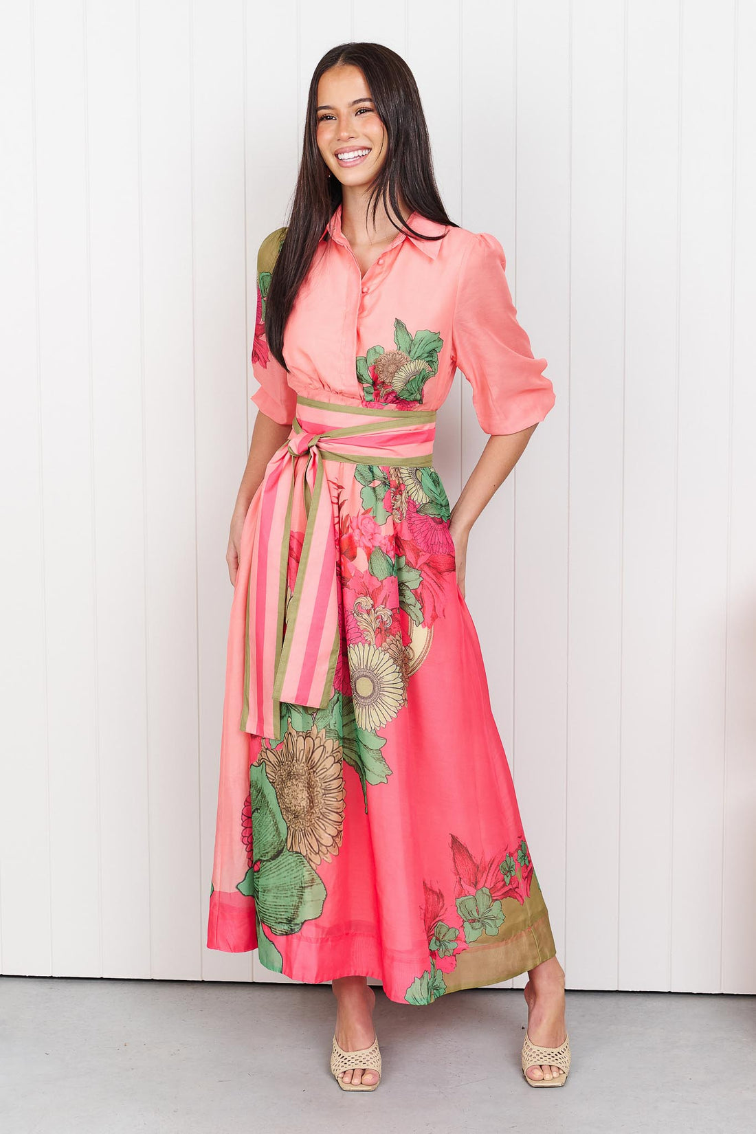 Woman wearing the minnie + MAXXIE Ayla Madison Ave midi event dress in peach and pink with a floral print and stripe waist tie, standing in front of a white panelled wall.