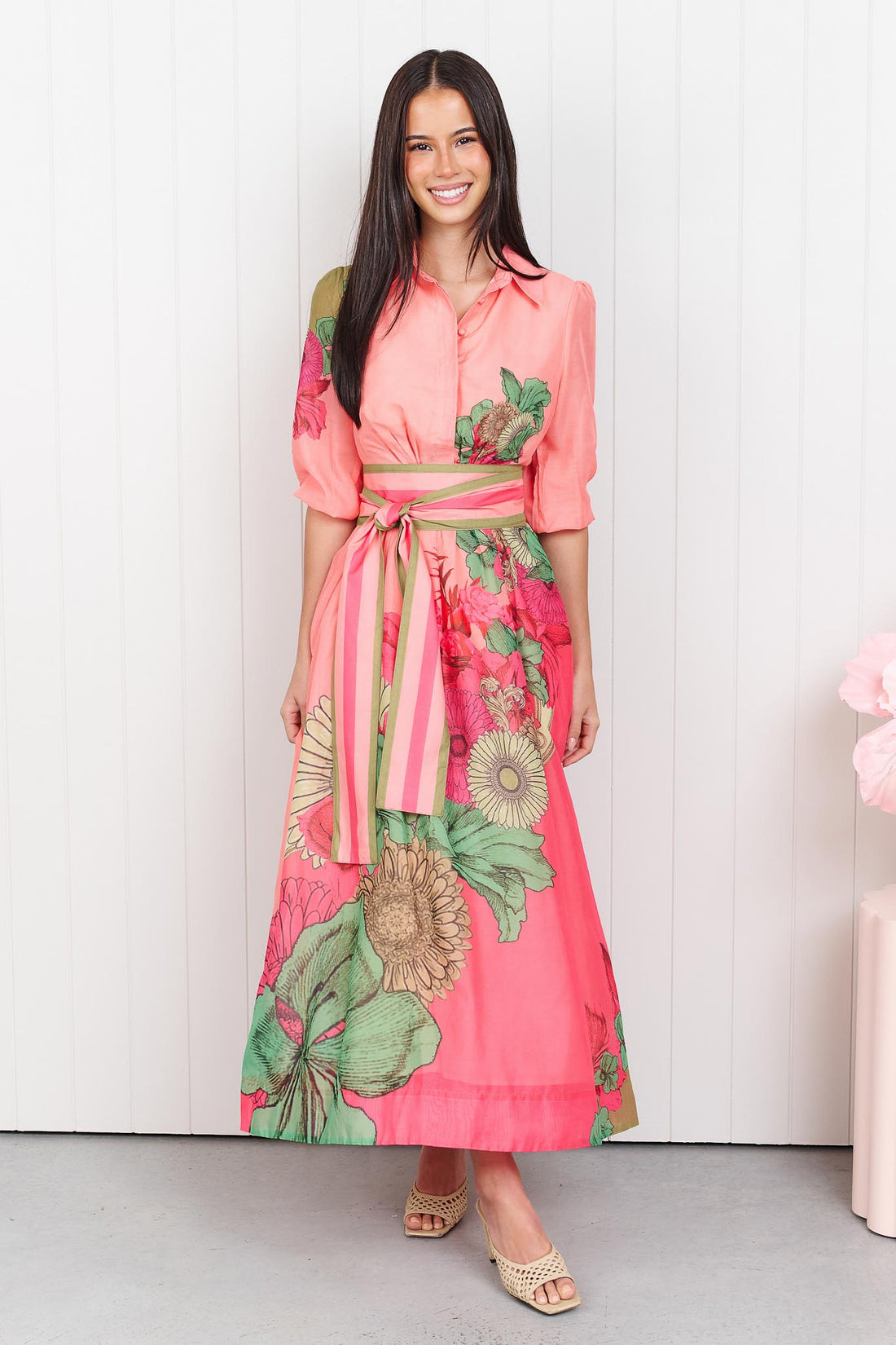 Woman wearing the minnie + MAXXIE Ayla Madison Ave midi event dress in peach and pink with a floral print and stripe waist tie, standing in front of a white panelled wall.