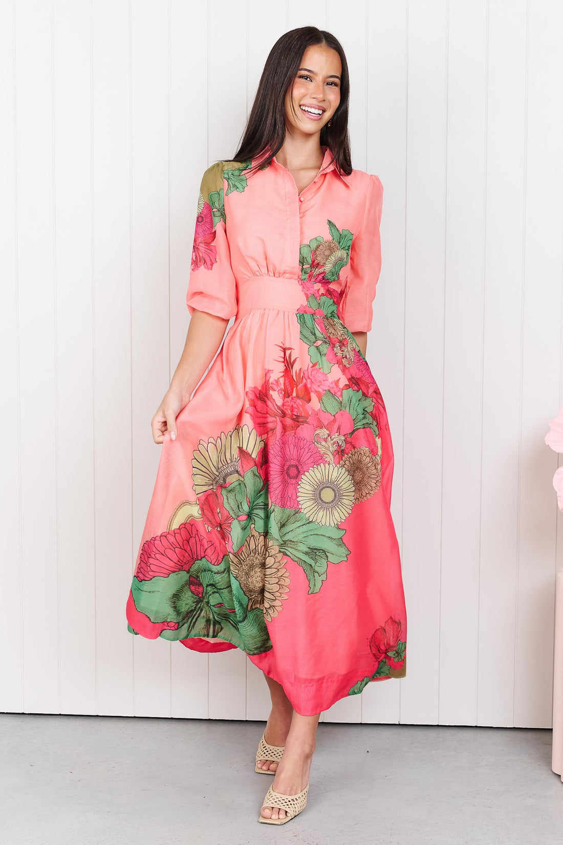 Woman wearing the minnie + MAXXIE Ayla Madison Ave midi event dress in peach and pink with a floral print, standing in front of a white panelled wall.