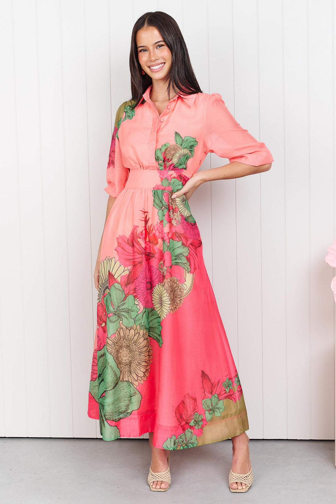 Woman wearing the minnie + MAXXIE Ayla Madison Ave midi event dress in peach and pink with a floral print, standing in front of a white panelled wall.