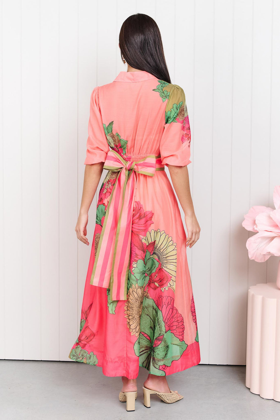 Back view of a woman wearing the minnie + MAXXIE Ayla Madison Ave midi event dress in peach and pink with a floral print and stripe waist tie bow, standing in front of a white panelled wall.